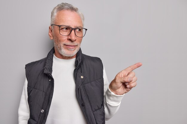 El hombre muestra una promoción presenta un nuevo producto con expresión segura de sí mismo usa gafas, un jersey informal y un chaleco muestra un espacio en blanco en gris