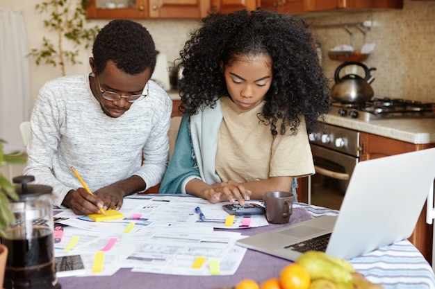 Hombre y mujer africanos sentados en la mesa de la cocina con papeles y computadora portátil, administrando las finanzas domésticas juntos: esposa contando con calculadora mientras el esposo toma notas con lápiz. Presupuesto familiar