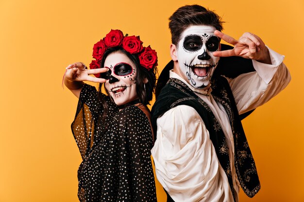 Hombre y mujer alegres se divierten en la pared naranja y muestran el signo de la paz. Retrato de pareja pintada en trajes nacionales mexicanos.