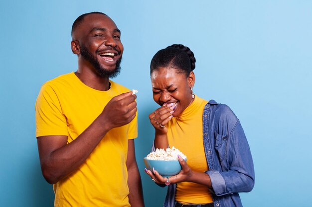 Hombre y mujer alegres viendo películas divertidas en la televisión, comiendo palomitas de maíz en el estudio y riendo. Pareja feliz con bocadillos mirando películas de comedia en la televisión, divirtiéndose y disfrutando de la risa juntos.