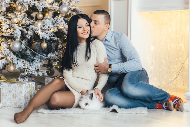Hombre y mujer bonita con un pequeño conejo posan ante un brillante árbol de Navidad