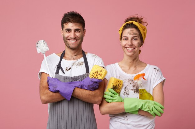 Hombre y mujer casados felices vistiendo ropa casual de pie con las manos cruzadas alegres de limpiar su casa con equipo de limpieza aislado