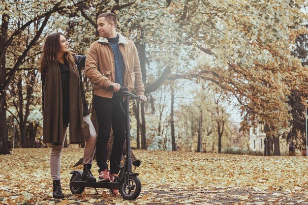 Un hombre y una mujer felices tienen un agradable paseo en scooter en el parque de otoño dorado.