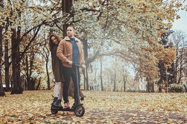 Un hombre y una mujer felices tienen un agradable paseo en scooter en el parque de otoño dorado.