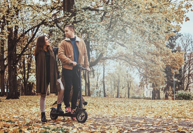 Un hombre y una mujer felices tienen un agradable paseo en scooter en el parque de otoño dorado.