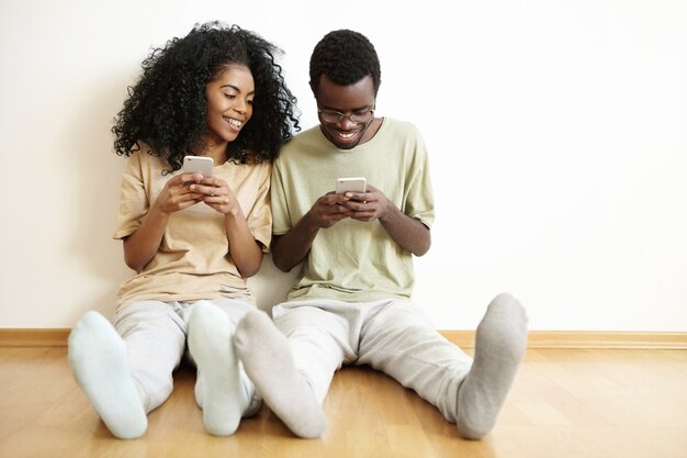 Hombre y mujer joven de piel oscura en ropa casual pasando tiempo juntos en el interior, jugando videojuegos en línea en dispositivos electrónicos