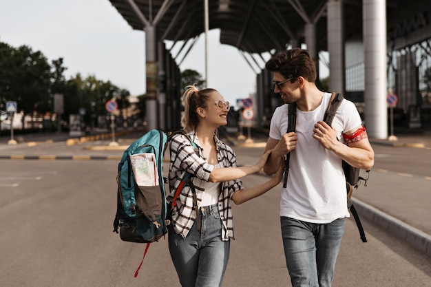 Hombre y mujer jóvenes en jeans y camisas blancas hablan y caminan cerca del aeropuerto Los viajeros con mochilas sonríen y se mueven
