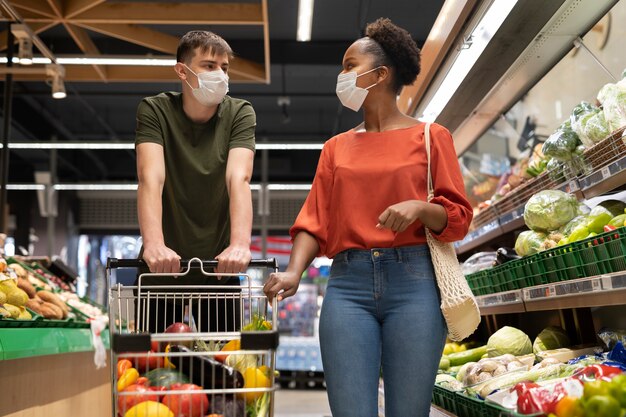 Hombre y mujer con máscaras médicas comprando comestibles con carrito de compras