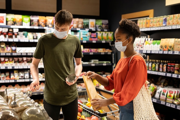 Hombre y mujer con máscaras médicas comprando comestibles con carrito de compras