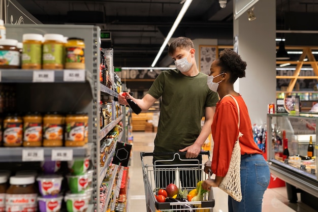 Hombre y mujer con máscaras médicas comprando comestibles con carrito de compras