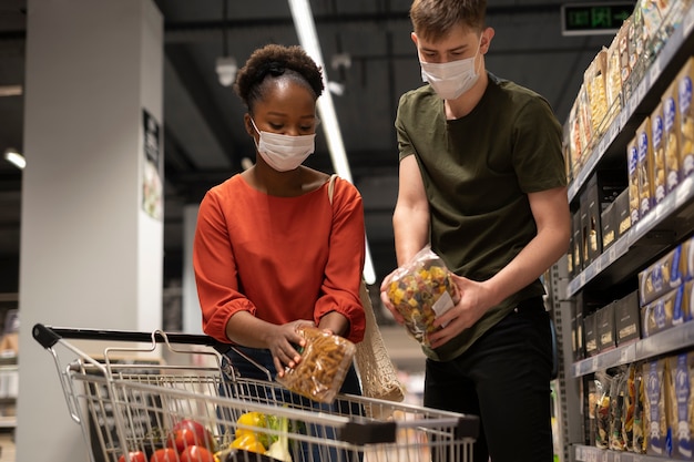 Hombre y mujer con máscaras médicas comprando comestibles con carrito de compras