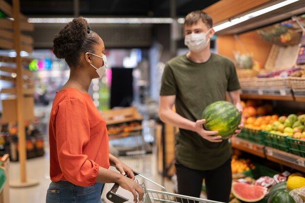 Hombre y mujer con máscaras médicas comprando comestibles con carrito de compras