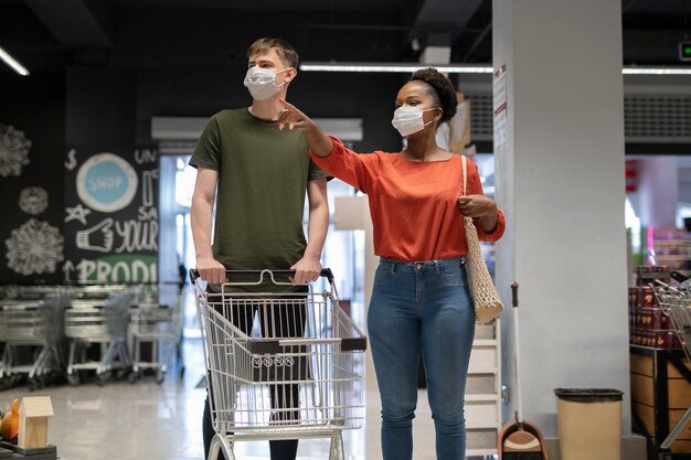 Hombre y mujer con máscaras médicas comprando comestibles con carrito de compras