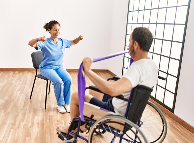 Hombre y mujer de mediana edad en sesión de rehabilitación usando banda elástica sentado en silla de ruedas en la clínica de fisioterapia