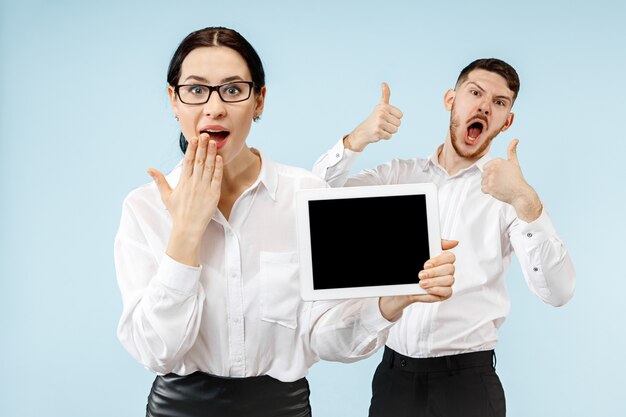 El hombre y la mujer de negocios sorprendidos sonriendo en una pared azul y mostrando la pantalla vacía de la computadora portátil o tableta