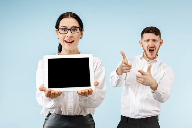 El hombre y la mujer de negocios sorprendidos sonriendo sobre un fondo de estudio azul y mostrando la pantalla vacía de la computadora portátil o tableta