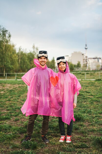 Hombre y mujer de pie en el campo con un impermeable de plástico rosa compartido y se quitaron los auriculares VR