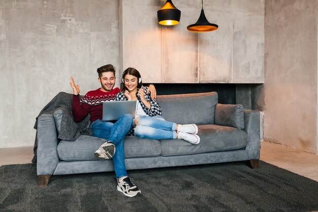 Hombre y mujer sonrientes jóvenes sentados en casa en invierno mirando en la computadora portátil con expresión de cara feliz, usando internet, pareja en el tiempo libre juntos, emoción positiva, citas