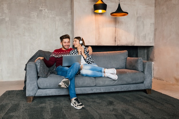 Foto gratuita hombre y mujer sonrientes jóvenes sentados en casa en invierno mirando en la computadora portátil con expresión de cara feliz, usando internet