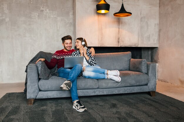 Hombre y mujer sonrientes jóvenes sentados en casa en invierno mirando en la computadora portátil con expresión de cara feliz, usando internet