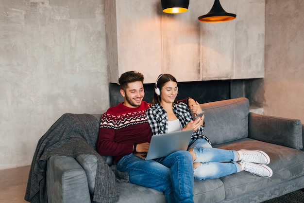 Hombre y mujer sonrientes jóvenes sentados en casa en invierno, trabajando en la computadora portátil, sosteniendo el teléfono inteligente, escuchando auriculares, pareja en el ocio pasando tiempo en línea, autónomo, citas