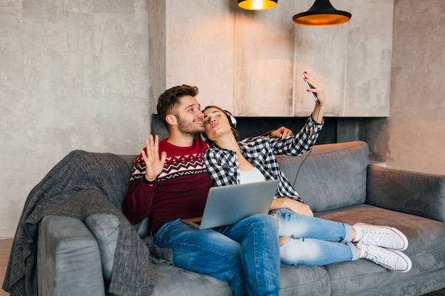 Hombre y mujer sonrientes jóvenes sentados en casa en invierno, trabajando en la computadora portátil, sosteniendo el teléfono inteligente, escuchando auriculares, pareja en el ocio pasando tiempo en línea, autónomo, citas