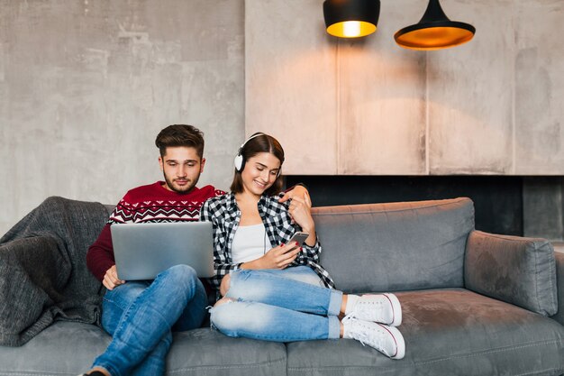 Hombre y mujer sonrientes jóvenes sentados en casa en invierno, trabajando en la computadora portátil, sosteniendo el teléfono inteligente, escuchando auriculares, pareja en el tiempo libre que pasa en línea, autónomo, citas