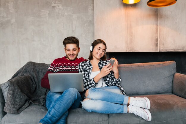 Hombre y mujer sonrientes jóvenes sentados en casa en invierno, trabajando en la computadora portátil, sosteniendo el teléfono inteligente, escuchando auriculares, pareja en el tiempo libre que pasa en línea, autónomo, citas