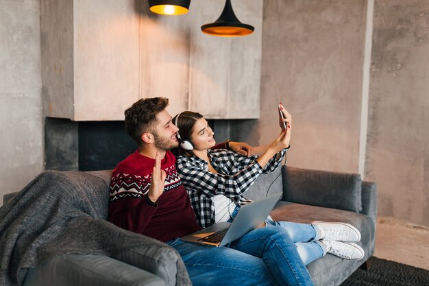 Hombre y mujer sonrientes jóvenes sentados en casa en invierno, trabajando en la computadora portátil, sosteniendo el teléfono inteligente, escuchando auriculares, pareja en el tiempo libre que pasa en línea, autónomo, citas