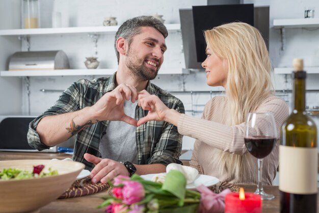 Hombre y mujer sonrientes que muestran el corazón por las manos y que se sientan en la tabla en cocina