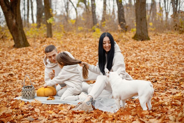 Hombre mujer su niña y perro sentados en una manta en el bosque de otoño Mujer morena y hombre juegan con su hija Familia vistiendo trajes deportivos beige