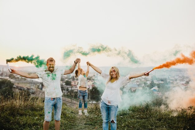 Hombre, mujer y su pequeña hija se divierten corriendo con humo colorido en sus brazos