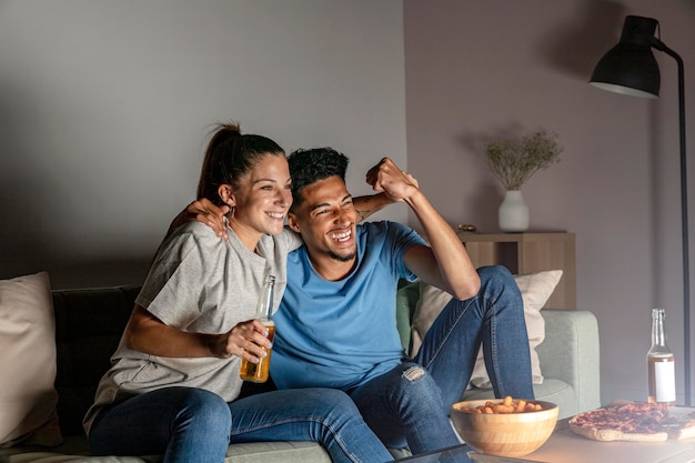 Hombre y mujer tomando cerveza en casa mientras ve la televisión y come bocadillos