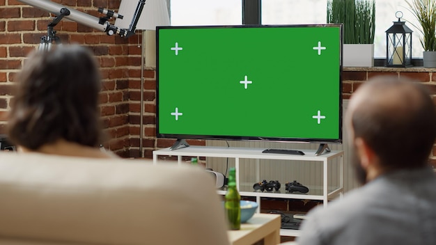 Hombre y mujer viendo la pantalla verde en la pantalla de televisión en la sala de estar. Personas que miran el espacio de copia aislado con plantilla de maqueta en blanco y fondo de clave de croma en la TV. Disparo de trípode. De cerca.