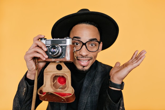 Hombre mulato de ojos marrones inspirado sosteniendo la cámara en la pared amarilla. Foto interior de primer plano del fotógrafo africano masculino disfrutando del trabajo.