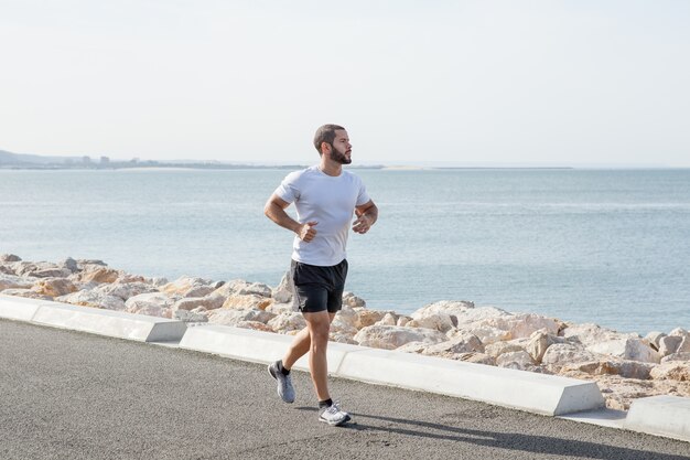 Hombre muscular serio corriendo en el camino de la playa