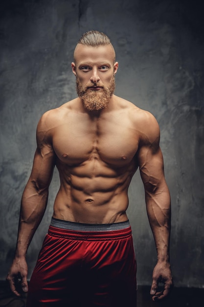 Hombre musculoso sin camisa con barba posando sobre fondo gris.