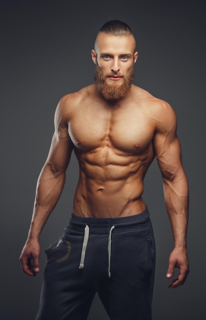 Hombre musculoso sin camisa con barba sobre un fondo gris.