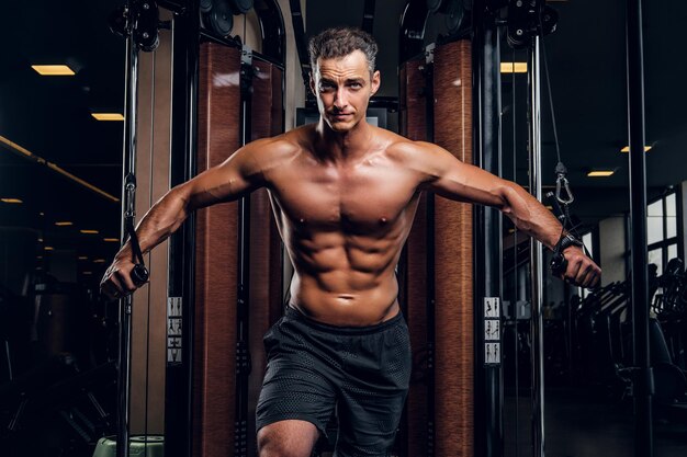 Un hombre musculoso sin camisa está haciendo ejercicios con aparatos de entrenamiento en un club de gimnasia oscuro.