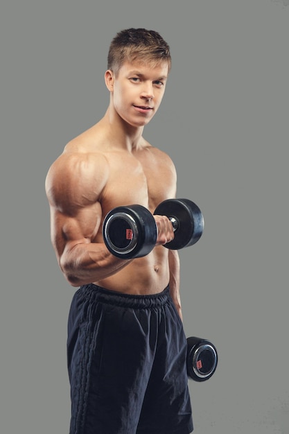 Hombre musculoso sin camisa haciendo ejercicios de bíceps con pesas sobre un fondo gris.