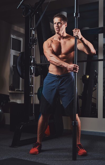 Un hombre musculoso sin camisa posando con una barra en el gimnasio.