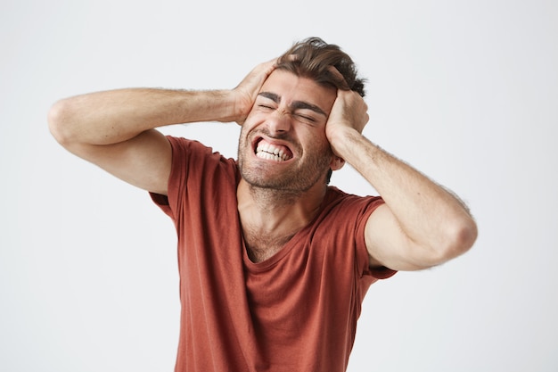 Hombre musculoso enojado emocional con camiseta roja cerrando los ojos con fuerza y gritando de dolor o incredulidad, manteniendo las manos sobre su cabeza. Emociones y sentimientos humanos negativos