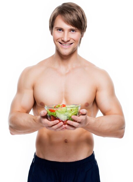 Hombre musculoso feliz joven que sostiene una ensalada sobre la pared blanca.