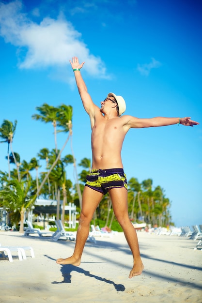 Hombre musculoso guapo feliz en sombrero para el sol en la playa saltando detrás del cielo azul detrás del cielo azul