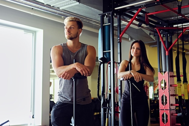 Hombre musculoso rubio y mujer morena atlética sostienen una barra en un club de gimnasia.