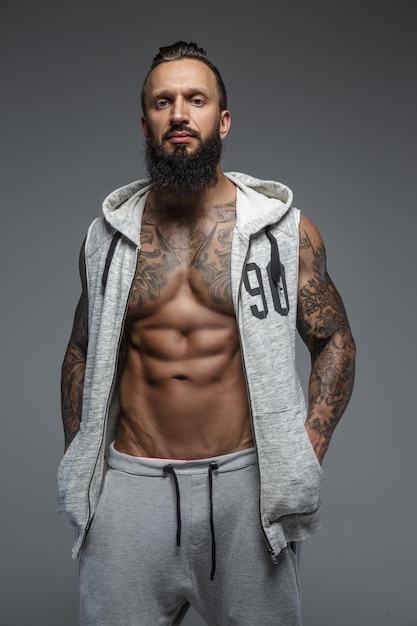 Hombre musculoso tatuado con barba en sudaderas grises zipuo. Aislado sobre fondo gris.
