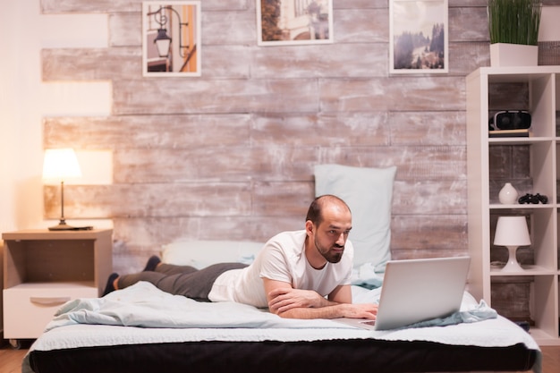 Hombre navegando en la computadora portátil por la noche en la cama en pijama.