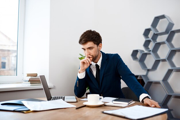 Hombre de negocios acertado joven que se sienta en el lugar de trabajo, fondo de la oficina.