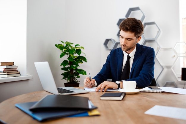 Hombre de negocios acertado joven que se sienta en el lugar de trabajo, fondo de la oficina.