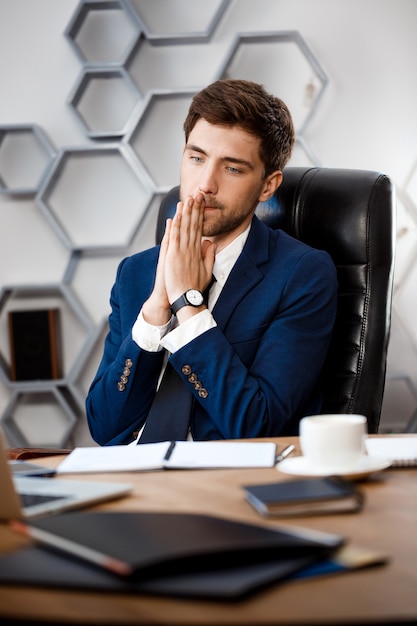 Foto gratuita hombre de negocios acertado joven que se sienta en el lugar de trabajo, fondo de la oficina.